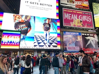 A bustling urban scene with large advertising billboards. The largest billboard is from American Eagle Outfitters, featuring models wearing t-shirts with 'Love is Love' text in rainbow colors, supporting the It Gets Better Project. Surrounding the main advertisement are other colorful billboards for shows, including 'Mean Girls' and 'Waitress'. A diverse crowd walks and takes photos below, adding to the vibrant city atmosphere.