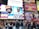 A bustling urban scene with large advertising billboards. The largest billboard is from American Eagle Outfitters, featuring models wearing t-shirts with 'Love is Love' text in rainbow colors, supporting the It Gets Better Project. Surrounding the main advertisement are other colorful billboards for shows, including 'Mean Girls' and 'Waitress'. A diverse crowd walks and takes photos below, adding to the vibrant city atmosphere.
