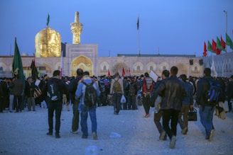 A large gathering of people near a religious site with an illuminated golden dome and minaret. The crowd appears to be engaged in an event or pilgrimage, with many individuals carrying backpacks and wearing various types of clothing. Flags of different colors are visible in the background, contributing to the ceremonial atmosphere.