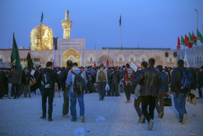 A large gathering of people near a religious site with an illuminated golden dome and minaret. The crowd appears to be engaged in an event or pilgrimage, with many individuals carrying backpacks and wearing various types of clothing. Flags of different colors are visible in the background, contributing to the ceremonial atmosphere.
