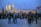 A large gathering of people near a religious site with an illuminated golden dome and minaret. The crowd appears to be engaged in an event or pilgrimage, with many individuals carrying backpacks and wearing various types of clothing. Flags of different colors are visible in the background, contributing to the ceremonial atmosphere.