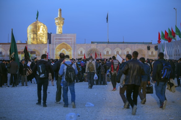 A large gathering of people near a religious site with an illuminated golden dome and minaret. The crowd appears to be engaged in an event or pilgrimage, with many individuals carrying backpacks and wearing various types of clothing. Flags of different colors are visible in the background, contributing to the ceremonial atmosphere.