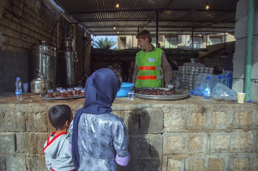 A street vendor wearing a green vest and red-striped shirt stands behind a makeshift counter, offering snacks and beverages. In front of him, trays with glasses of tea are arranged alongside a large dish filled with dates. Two children, one in a hijab and the other in a red-striped shirt, observe the vendor. The background reveals stacks of packaged goods and several water bottles, indicating a bustling market atmosphere.