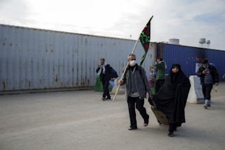 A group of people is walking near large, worn shipping containers. The individuals are dressed in casual and modest clothing, with some wearing face masks. One person holds a flag with intricate designs, and another carries a suitcase. The background shows cloudy skies.