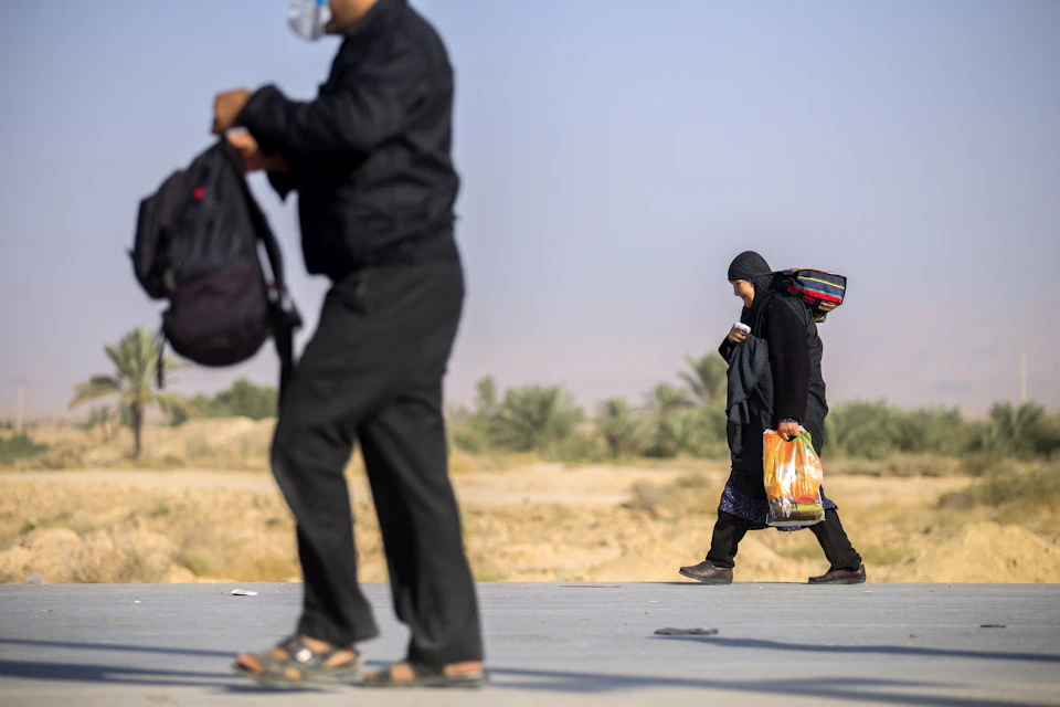 The image depicts two individuals walking on a road, each carrying bags and backpacks. The person on the left is wearing a black jacket and has a backpack slung over their shoulder. They are also holding a handkerchief to their face. On the right side of the road, another individual can be seen, dressed in an orange shirt and carrying a bag with them.

The background features a desert landscape with palm trees scattered across it, suggesting that they might be walking through a desert region or near one. The sky is clear blue, indicating good weather conditions for their journey.