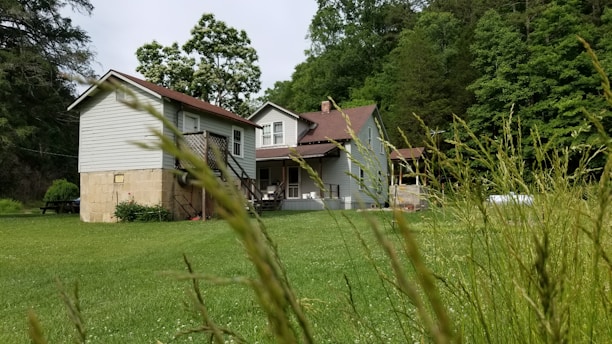A rustic house with a red roof is surrounded by lush greenery, tall grass, and trees. The architecture suggests a countryside setting, and there's a picnic table located under the shade of tall trees, creating a serene atmosphere.