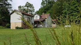 A rustic house with a red roof is surrounded by lush greenery, tall grass, and trees. The architecture suggests a countryside setting, and there's a picnic table located under the shade of tall trees, creating a serene atmosphere.