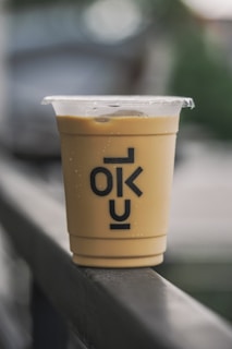 A plastic cup filled with a creamy, light brown beverage is placed on a railing. The cup has a clear plastic lid on top and the letters 'OKU' printed in bold black text on its side. The background is blurred, suggesting an outdoor setting.