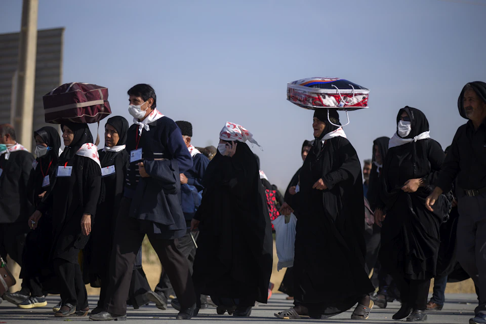 The image depicts a group of people walking down a street, each carrying bags and wearing traditional clothing. The individuals are dressed in black attire with white headscarves, suggesting they may be part of a cultural or religious community. Some of them have their heads covered by hats or scarves as well. The scene is set against an urban backdrop, including buildings and trees that add depth to the image.