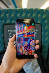 A hand holds a Samsung smartphone displaying colorful folk art. The art features whimsical characters, a bird, and abstract shapes in vibrant colors. The background includes the seat and interior of a train or bus, with a patterned blue-green fabric upholstery.
