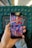 A hand holds a Samsung smartphone displaying colorful folk art. The art features whimsical characters, a bird, and abstract shapes in vibrant colors. The background includes the seat and interior of a train or bus, with a patterned blue-green fabric upholstery.
