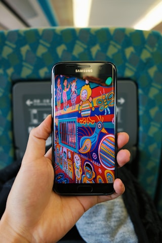 A hand holds a Samsung smartphone displaying colorful folk art. The art features whimsical characters, a bird, and abstract shapes in vibrant colors. The background includes the seat and interior of a train or bus, with a patterned blue-green fabric upholstery.
