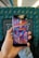 A hand holds a Samsung smartphone displaying colorful folk art. The art features whimsical characters, a bird, and abstract shapes in vibrant colors. The background includes the seat and interior of a train or bus, with a patterned blue-green fabric upholstery.