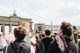 A group of people are gathered, facing towards a historic landmark with a sign that reads 'Wake up' and shows a picture of the Earth among flames. Some people are holding signs and some wear backpacks and jackets.