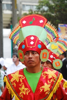 A person is dressed in colorful traditional attire featuring vibrant reds, greens, and yellows. The costume includes an intricate headdress decorated with geometric patterns, flowers, and symbols. The expression is serious and focused, while other individuals in similar outfits are visible in the background.