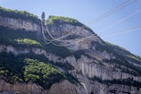 A high cliff with dense vegetation at the top is traversed by several cables in the sky. A structure is present at the top of the cliff, possibly a cable car or zip line station. The rugged cliff face displays layers of rock formations, and sunlight highlights sections of the greenery.