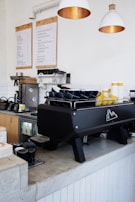 A modern coffee shop counter with a large espresso machine in black color. There are stacks of blue and yellow mugs on top of the machine. Wooden cabinets and a menu board are visible in the background, along with hanging white pendant lights.