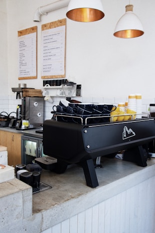 A modern coffee shop counter with a large espresso machine in black color. There are stacks of blue and yellow mugs on top of the machine. Wooden cabinets and a menu board are visible in the background, along with hanging white pendant lights.