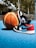A pair of black, white, and red sneakers is placed next to a basketball on a blue outdoor court. The background features blurred trees and a structure with stairs.