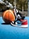 A pair of black, white, and red sneakers is placed next to a basketball on a blue outdoor court. The background features blurred trees and a structure with stairs.