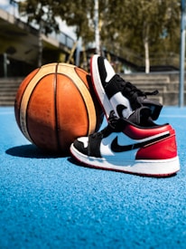 A pair of black, white, and red sneakers is placed next to a basketball on a blue outdoor court. The background features blurred trees and a structure with stairs.