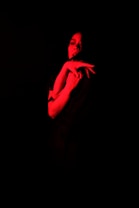 A person is illuminated by a deep red light, creating a dramatic and mysterious atmosphere. The background is almost entirely black, adding to the contrast and focus on the subject's expression and posture.