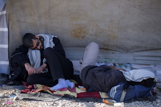 Two people are resting on the ground under a makeshift shelter. One is sitting upright, while the other is lying down with a cloth over the head. They appear to be wearing casual clothing and are using a blanket for comfort.