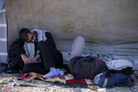 Two people are resting on the ground under a makeshift shelter. One is sitting upright, while the other is lying down with a cloth over the head. They appear to be wearing casual clothing and are using a blanket for comfort.