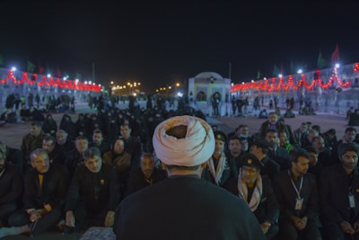 A large gathering of people seated in rows under the night sky. The central figure, seen from behind, wears a white turban, indicating a possible religious or cultural leader. The scene is illuminated by strings of red lights and distant streetlights, creating a festive atmosphere. Many attendees appear to be dressed in dark or traditional clothing, with some wearing badges or identification around their necks.