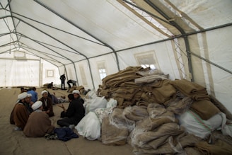 A large, open tent is filled with people sitting and interacting. Piles of folded blankets and bags are stacked to one side. The atmosphere appears communal and informal.