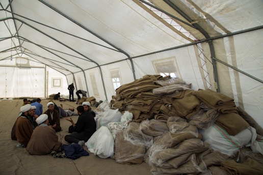 A large, open tent is filled with people sitting and interacting. Piles of folded blankets and bags are stacked to one side. The atmosphere appears communal and informal.