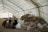 A large, open tent is filled with people sitting and interacting. Piles of folded blankets and bags are stacked to one side. The atmosphere appears communal and informal.