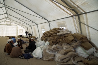 A large, open tent is filled with people sitting and interacting. Piles of folded blankets and bags are stacked to one side. The atmosphere appears communal and informal.