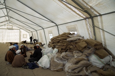 A large, open tent is filled with people sitting and interacting. Piles of folded blankets and bags are stacked to one side. The atmosphere appears communal and informal.