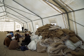 A large, open tent is filled with people sitting and interacting. Piles of folded blankets and bags are stacked to one side. The atmosphere appears communal and informal.