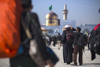 A large golden dome and minaret are prominent in the background, indicating a significant religious or cultural site. People are walking towards the building, some carrying backpacks. A person in the foreground adjusts another person’s headwear.