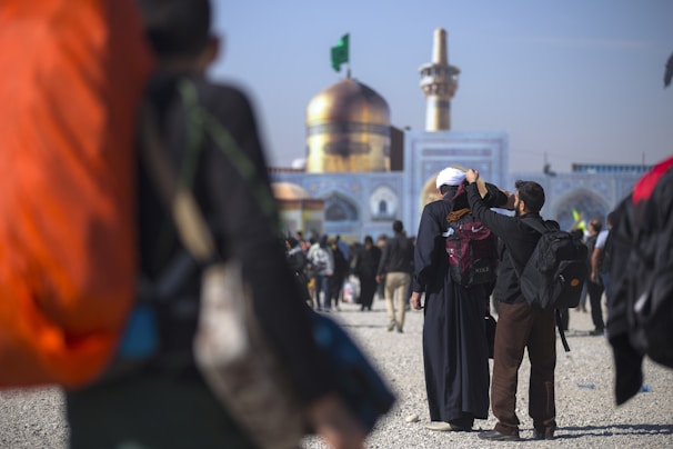 A large golden dome and minaret are prominent in the background, indicating a significant religious or cultural site. People are walking towards the building, some carrying backpacks. A person in the foreground adjusts another person’s headwear.