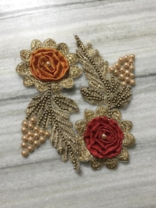 Two decorative floral motifs with intricate metallic embroidery against a light marble background. Each flower has petals made of gold-thread, accentuated by clusters of pearls. The left flower is orange with a central pearl, while the right flower is red, bringing a vibrant contrast.