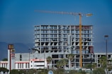 A large, unfinished concrete building structure is set against a clear blue sky. A yellow construction crane is prominently positioned next to the building, indicating ongoing construction activity. Nearby, palm trees and commercial signs, including logos for notable businesses, are visible. The background features a range of mountains providing a scenic backdrop.