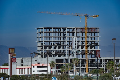 A large, unfinished concrete building structure is set against a clear blue sky. A yellow construction crane is prominently positioned next to the building, indicating ongoing construction activity. Nearby, palm trees and commercial signs, including logos for notable businesses, are visible. The background features a range of mountains providing a scenic backdrop.