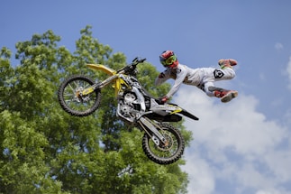 A motocross rider in the air performing a stunt with one hand on the handlebars and legs extended outwards from the motorcycle. The rider is wearing a white outfit with red and black boots, and a red helmet with goggles. The background features green foliage and a clear blue sky with a few clouds.