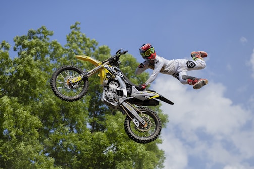 A motocross rider in the air performing a stunt with one hand on the handlebars and legs extended outwards from the motorcycle. The rider is wearing a white outfit with red and black boots, and a red helmet with goggles. The background features green foliage and a clear blue sky with a few clouds.