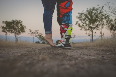 A pair of rugged motocross boots standing on rocky terrain with a blurred motorcycle in the background.