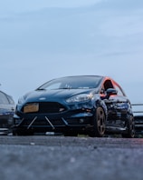 A black sports car with a New York license plate parked on asphalt, viewed from a low angle. The car has noticeable details like an aerodynamic front bumper and tinted windows. It is positioned next to another vehicle, with a blue sky in the background.
