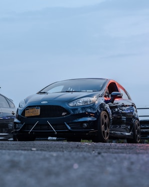 A black sports car with a New York license plate parked on asphalt, viewed from a low angle. The car has noticeable details like an aerodynamic front bumper and tinted windows. It is positioned next to another vehicle, with a blue sky in the background.