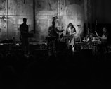 A black and white image of a live band performance with musicians playing instruments such as guitar, saxophone, double bass, and drums. The backdrop features abstract art and text, and the dimly lit stage creates silhouettes of the performers.