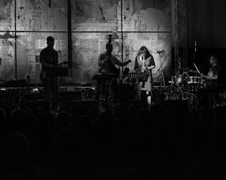 A black and white image of a live band performance with musicians playing instruments such as guitar, saxophone, double bass, and drums. The backdrop features abstract art and text, and the dimly lit stage creates silhouettes of the performers.