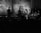 A black and white image of a live band performance with musicians playing instruments such as guitar, saxophone, double bass, and drums. The backdrop features abstract art and text, and the dimly lit stage creates silhouettes of the performers.