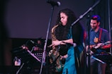 A musician is passionately playing a saxophone on stage, surrounded by other performers. The environment suggests a live music performance with microphones, music stands, and another musician playing a guitar, who is smiling and looking at the saxophonist.