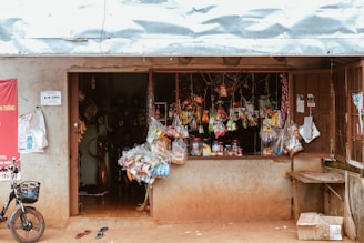 A small rustic shop with an open window display featuring a variety of hanging snacks and goods. The exterior walls are worn and weathered. A bicycle is parked in front, and a broom lies nearby. The interior is dimly lit, with various items stored inside.